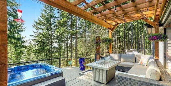 A wood-decked patio with a hot tub, a fire-pit coffee table, and comfortable sectional seating, overlooking a forest.