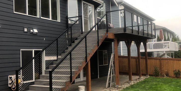 A backyard wooden deck with stairs and modern black metal railings attached to the back of a dark blue house.
