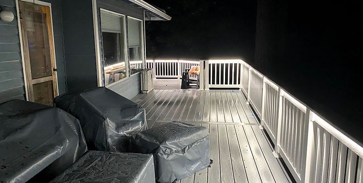 A gray wooden deck at night, illuminated by LED strip lights along the white railing and house exterior.