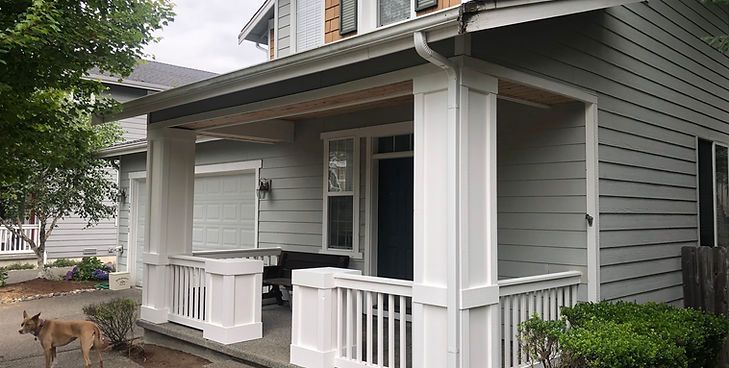 A gray, two-story house with a white-railed front porch, a garage, and a dog standing on the driveway.