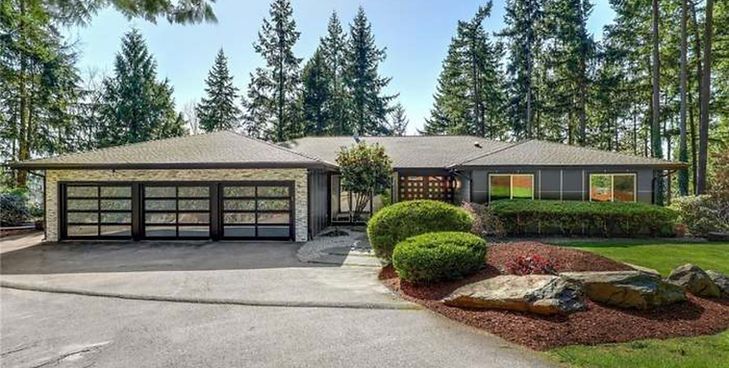 A single-story, gray house with a three-car glass garage, stone siding, and surrounding evergreen trees.