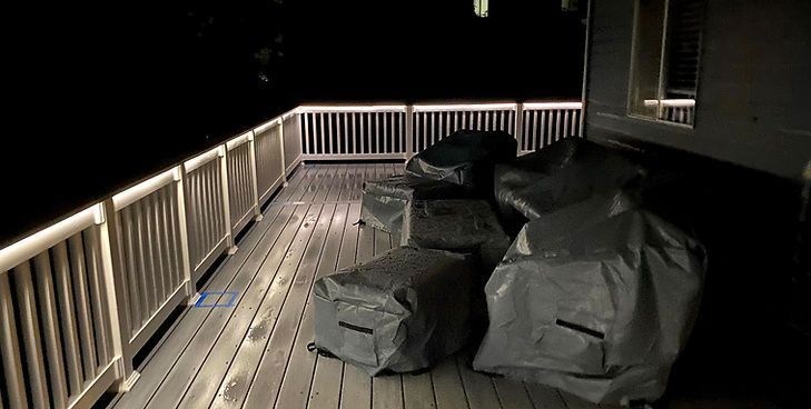 A dimly lit outdoor deck at night with furniture covered by grey protective tarps and illuminated railings.