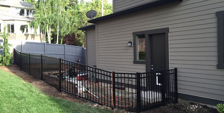 A black metal fence encloses a patio area next to the gray siding of a house with a back door.