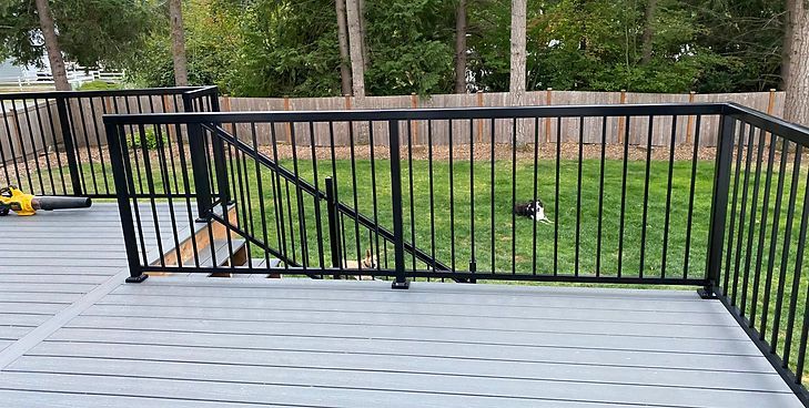 A grey composite deck features black metal railings, including a diagonal section descending to stairs overlooking a yard.