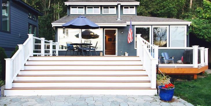A wide wooden deck with a large set of stairs leads up to a blue house with a patio table, umbrella, and American flag.