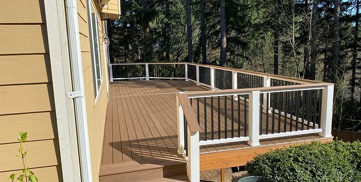 A wooden deck with a white railing and black balusters extends from a tan house, surrounded by trees.