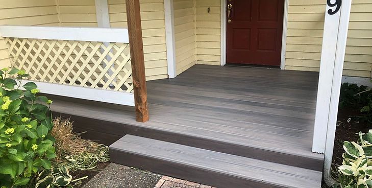 A front porch with grey composite decking, a single step, a white lattice railing, and a red front door on a yellow house.