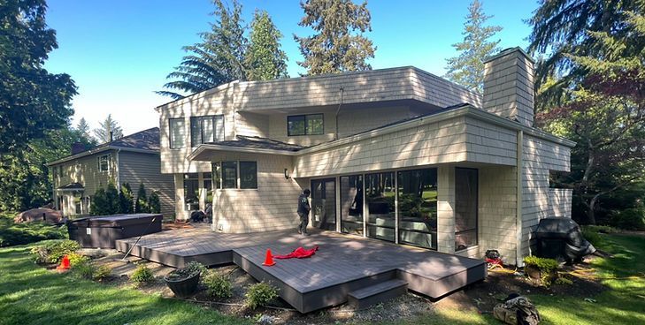 A beige two-story house with a wooden deck, a brick chimney, and large windows on a sunny day with trees in the background.