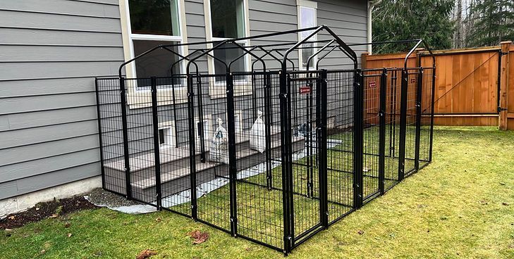 A black metal outdoor pet kennel enclosure attached to the exterior wall of a grey house.