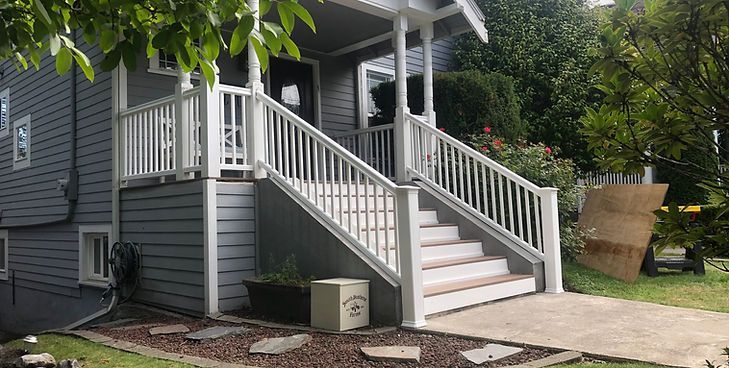 A gray house exterior with white railings on a front porch staircase leading to a concrete walkway.