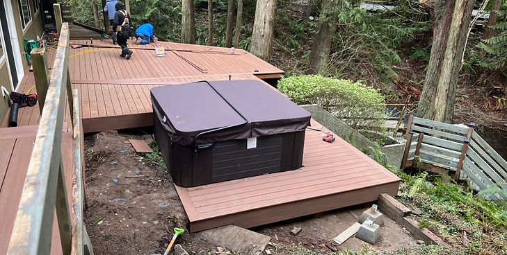 A dark brown hot tub sits on a new, reddish-brown composite deck in a forested yard next to wooden stairs.