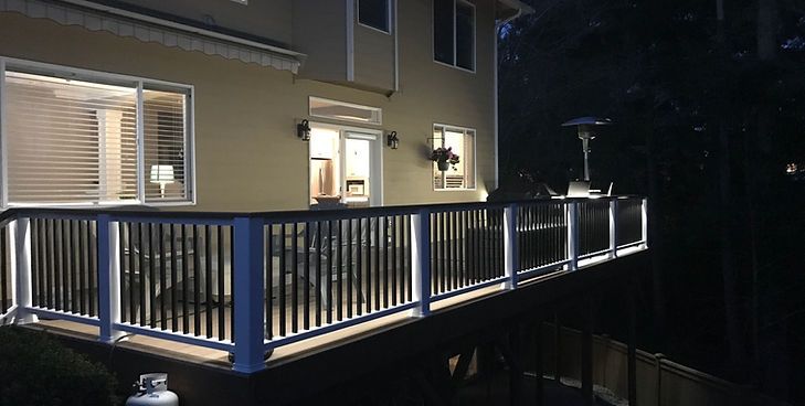 A two-story house exterior at night featuring a large wooden deck with illuminated white railings and patio lighting.