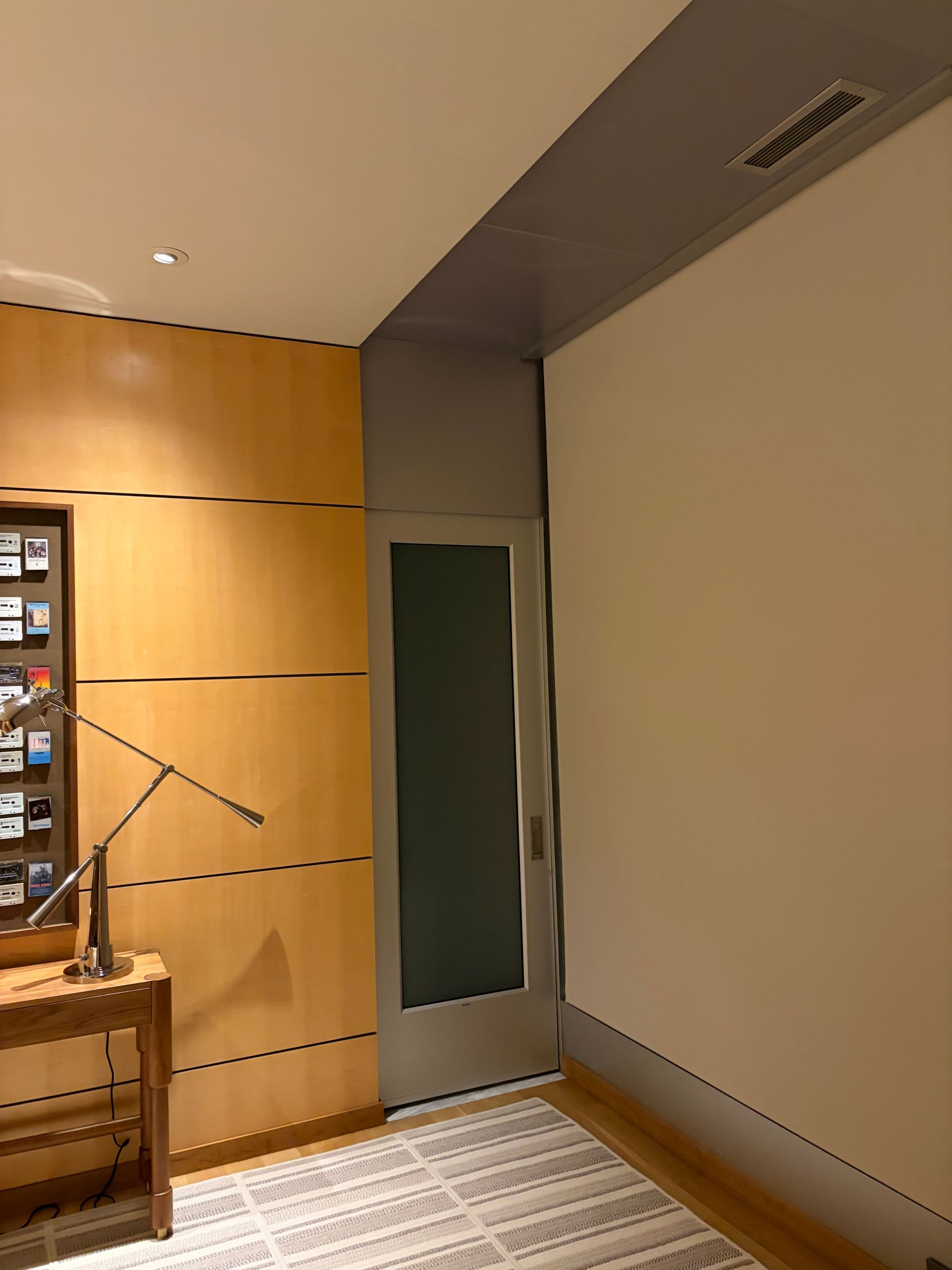 A view of a modern hallway with a wood-paneled wall, a frosted glass door, and a table with a lamp on the left.