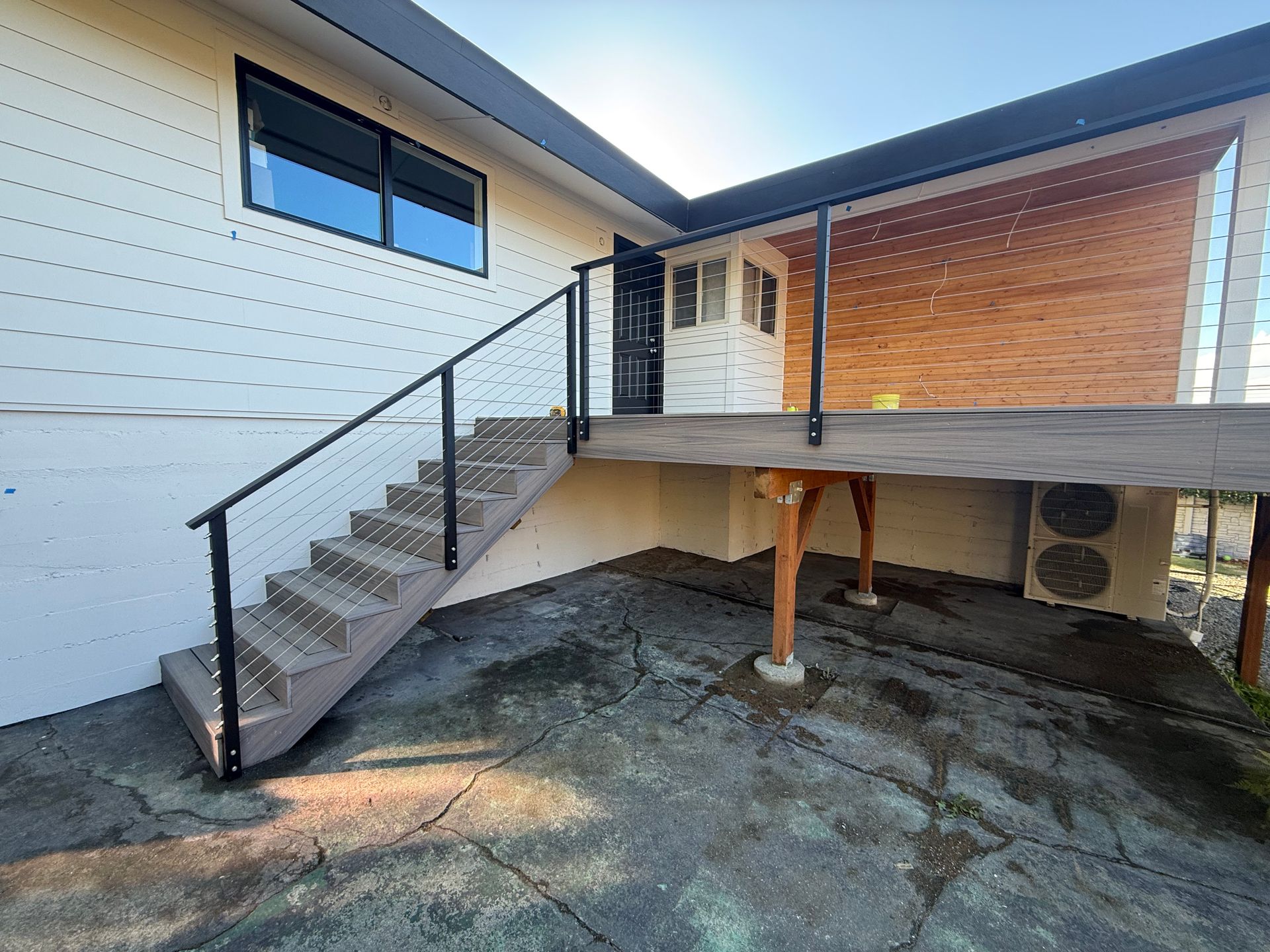 A raised wooden deck with a staircase featuring a black railing, set against a white building with wood-paneled accents.