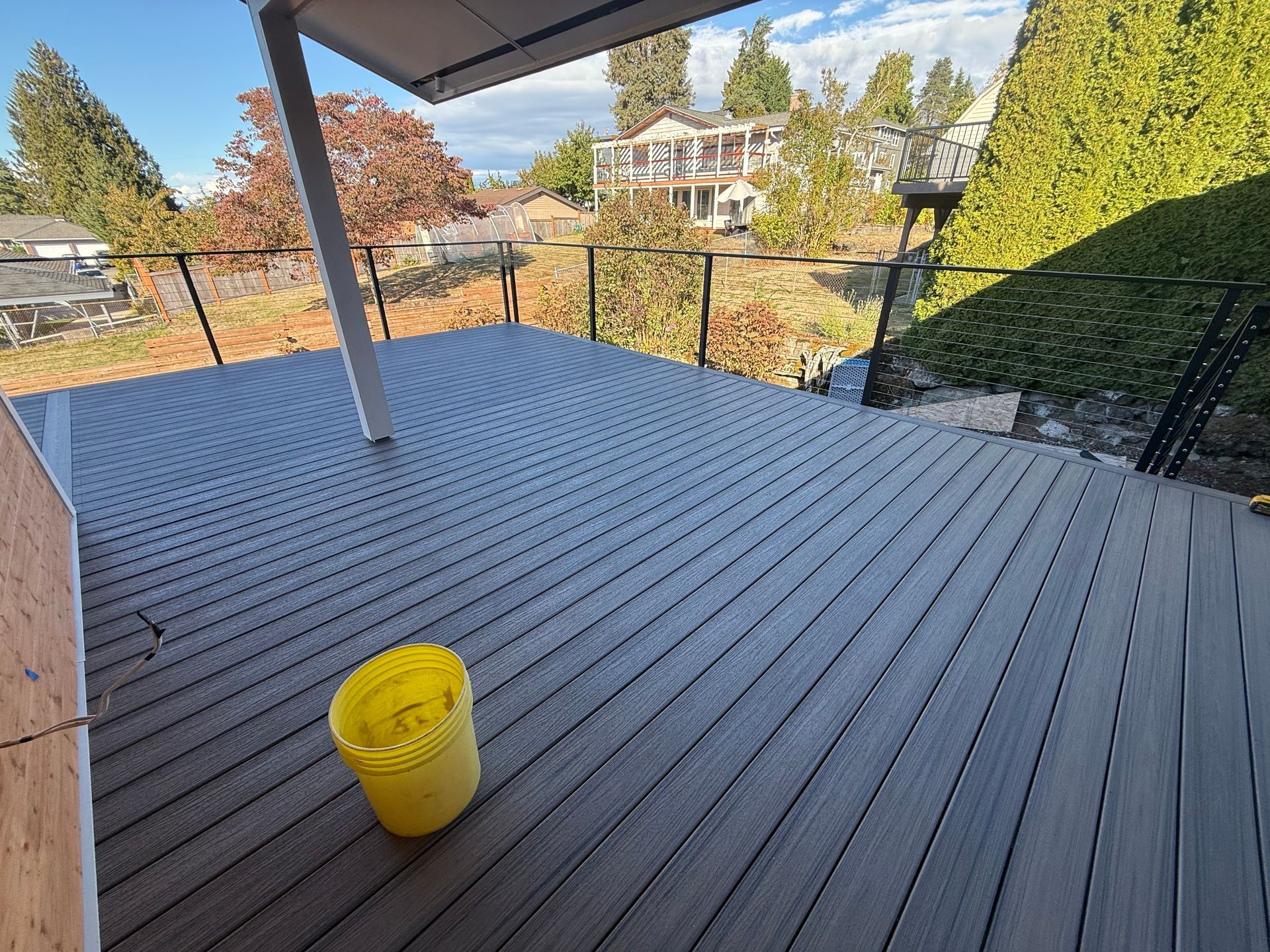 A modern grey composite deck with glass railings, a white support pillar, and a yellow bucket in a sunny outdoor setting.