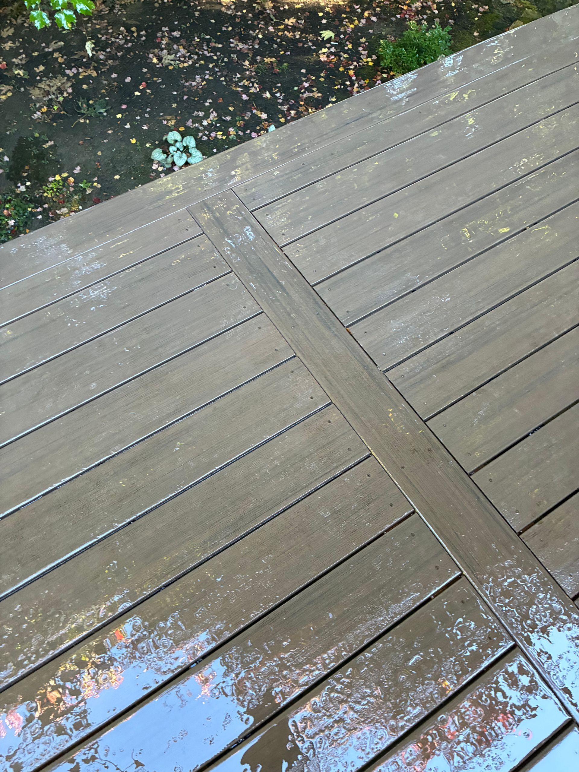 A rain-slicked wooden deck with parallel planks, viewed from a high angle, with scattered autumn leaves on the ground.