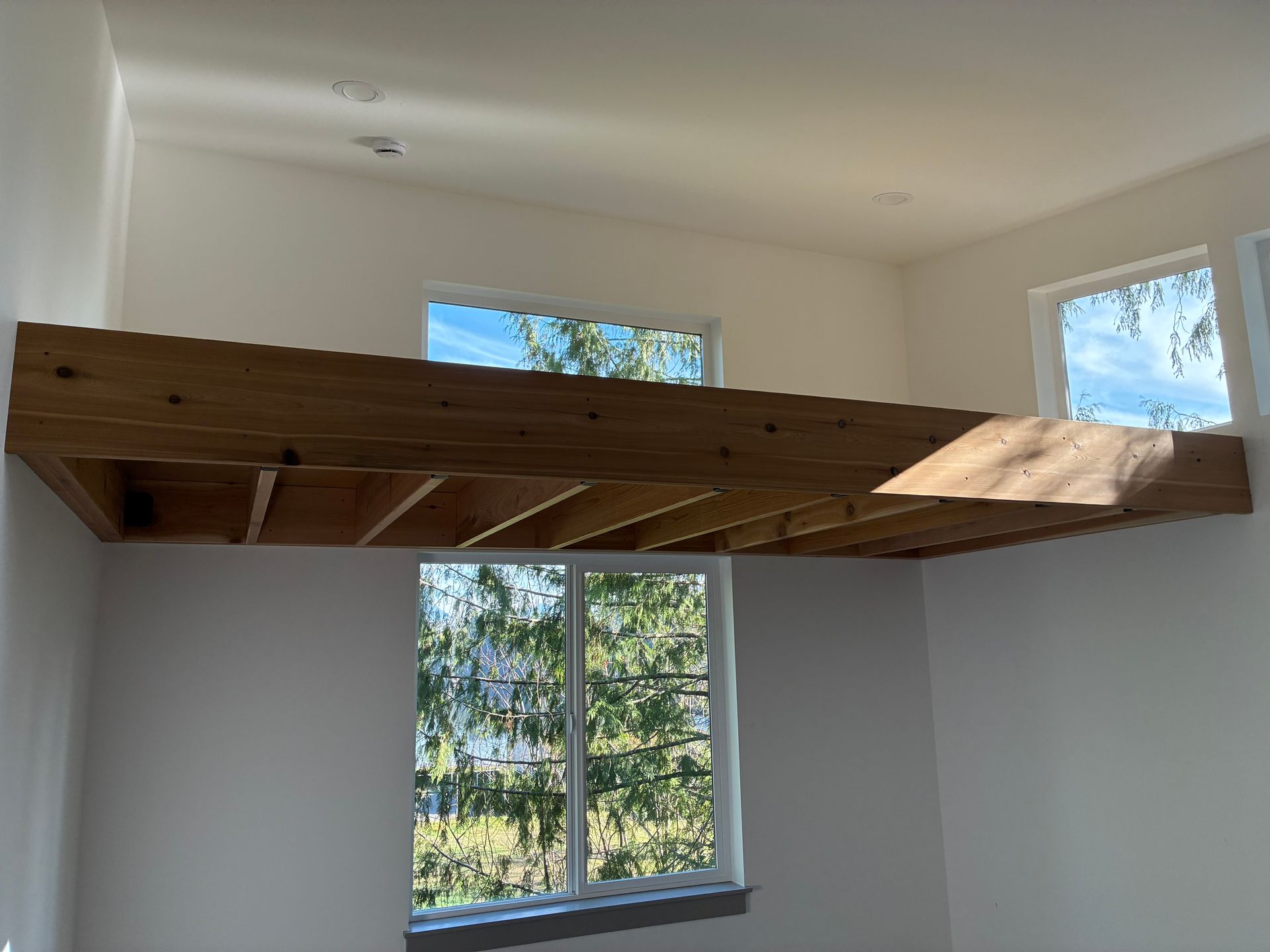 A wooden loft platform cantilevered from a white wall, positioned above a window looking out onto trees.