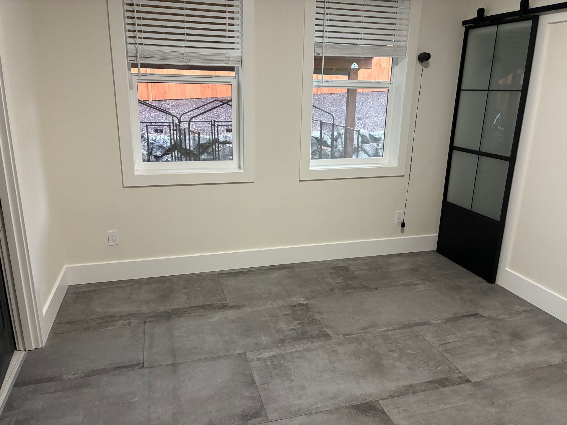 An empty room with light walls, large gray tile flooring, two windows with blinds, and a black framed sliding glass door.
