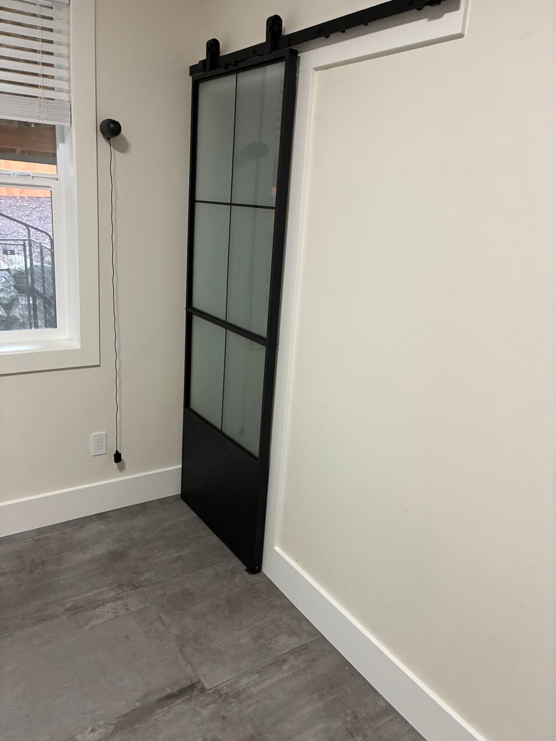 A black-framed barn door with frosted glass panels hangs on a track against a white wall next to a window.