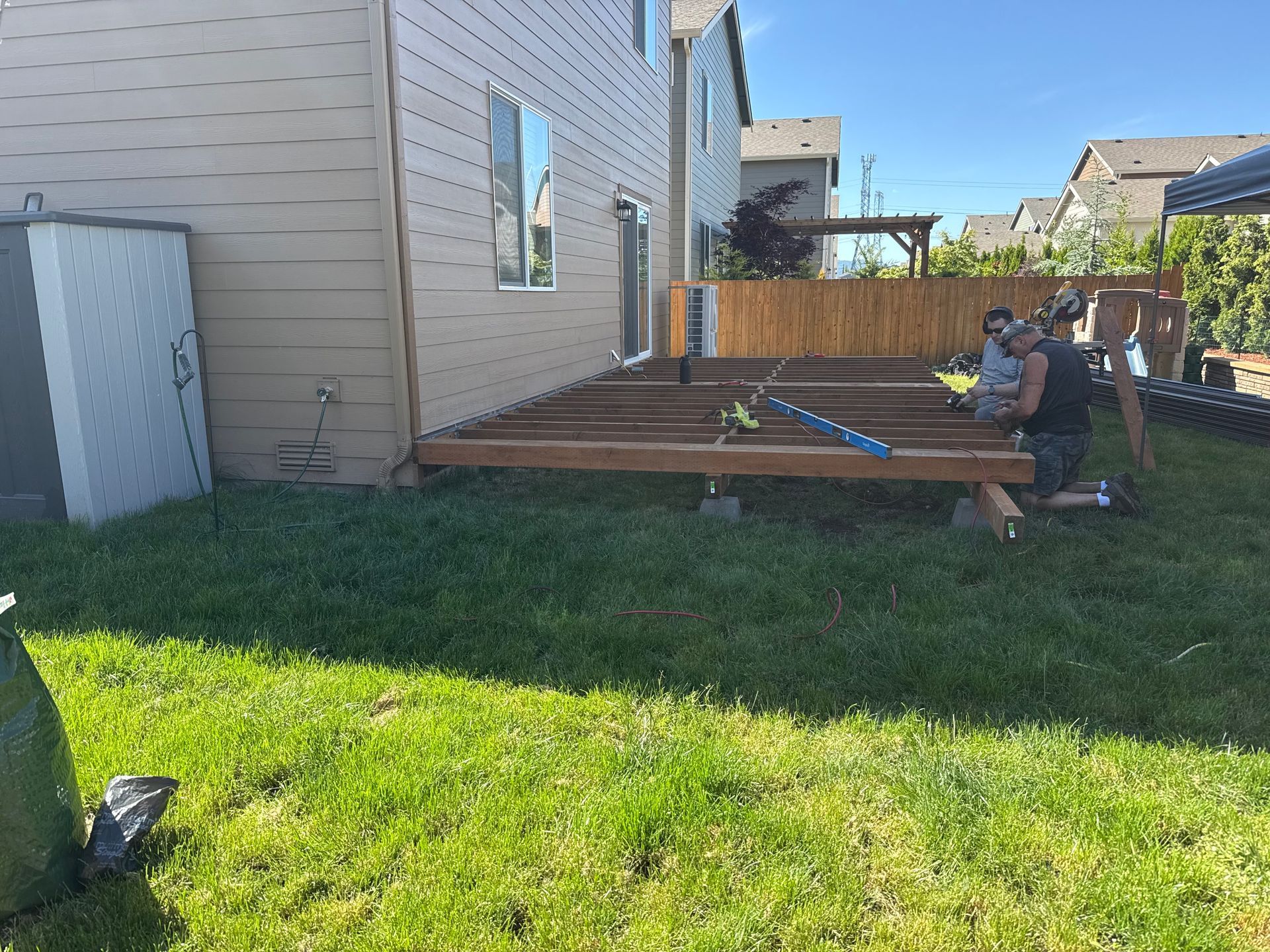 Two people work on the wooden framing of a backyard deck attached to the side of a beige house.