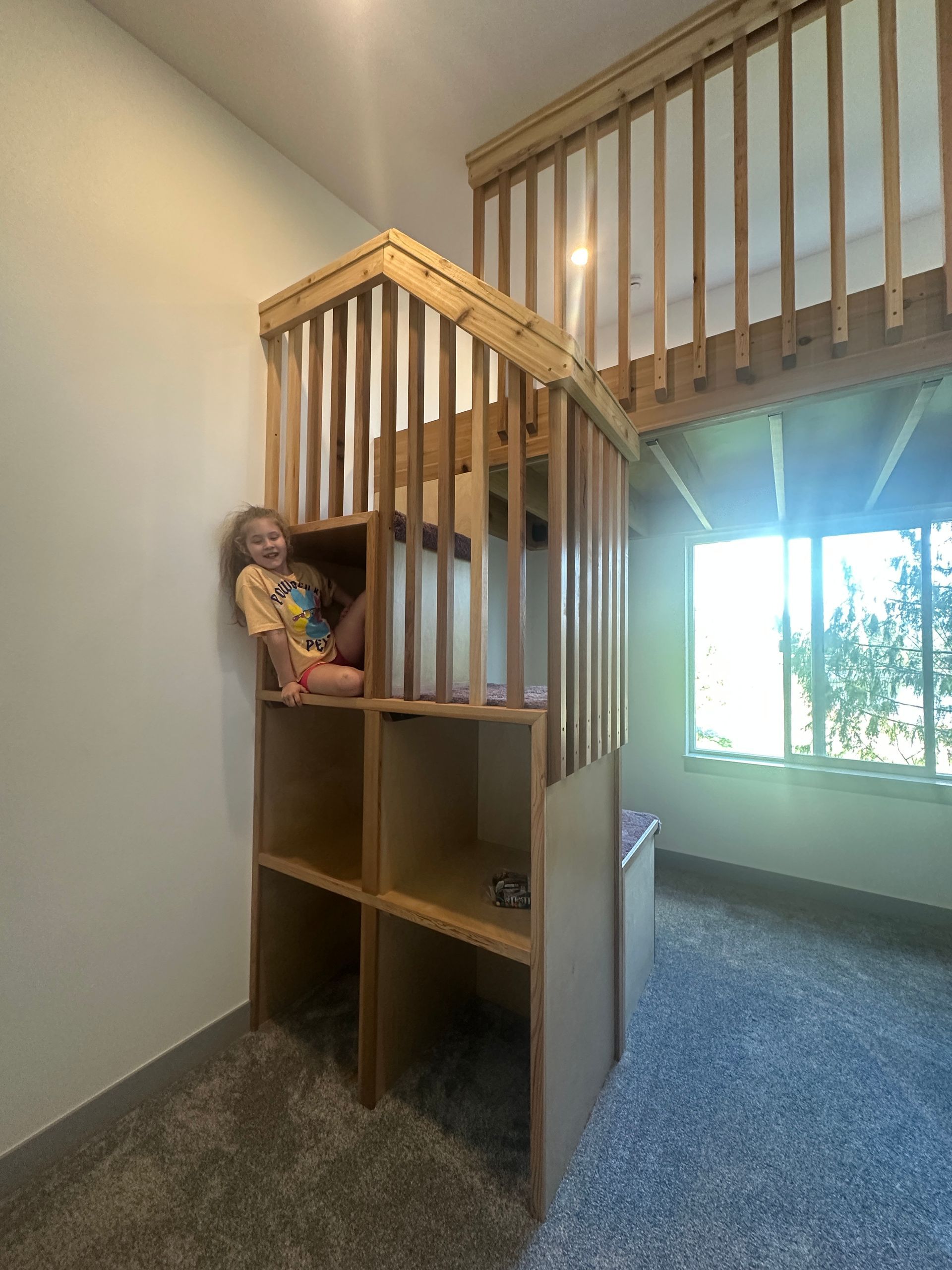 A child sits inside the middle cubby of a wooden loft bed structure featuring an open staircase and safety railing.