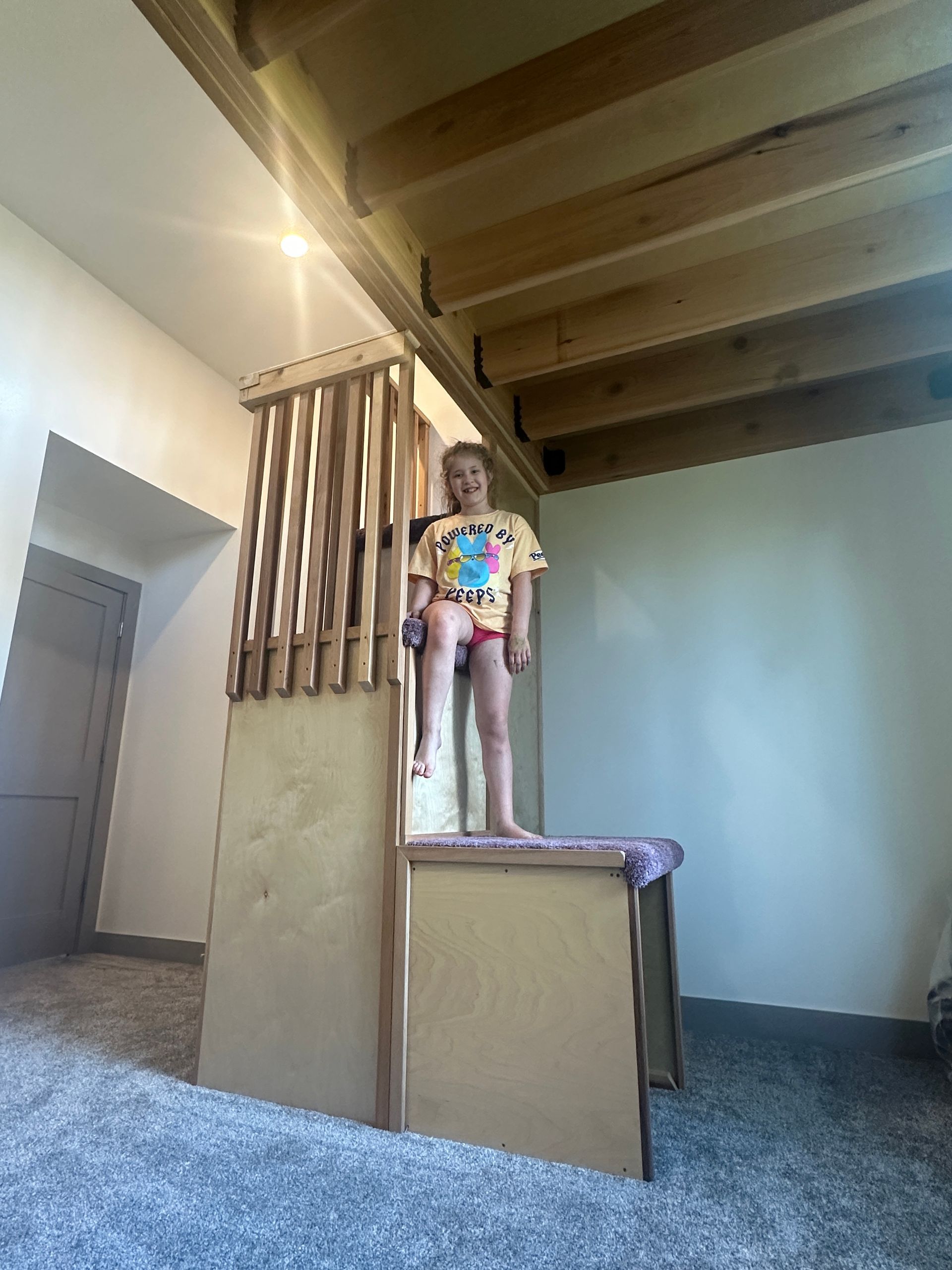 A person stands on a wooden platform structure, partially enclosed by vertical slats, in a room with exposed ceiling beams.