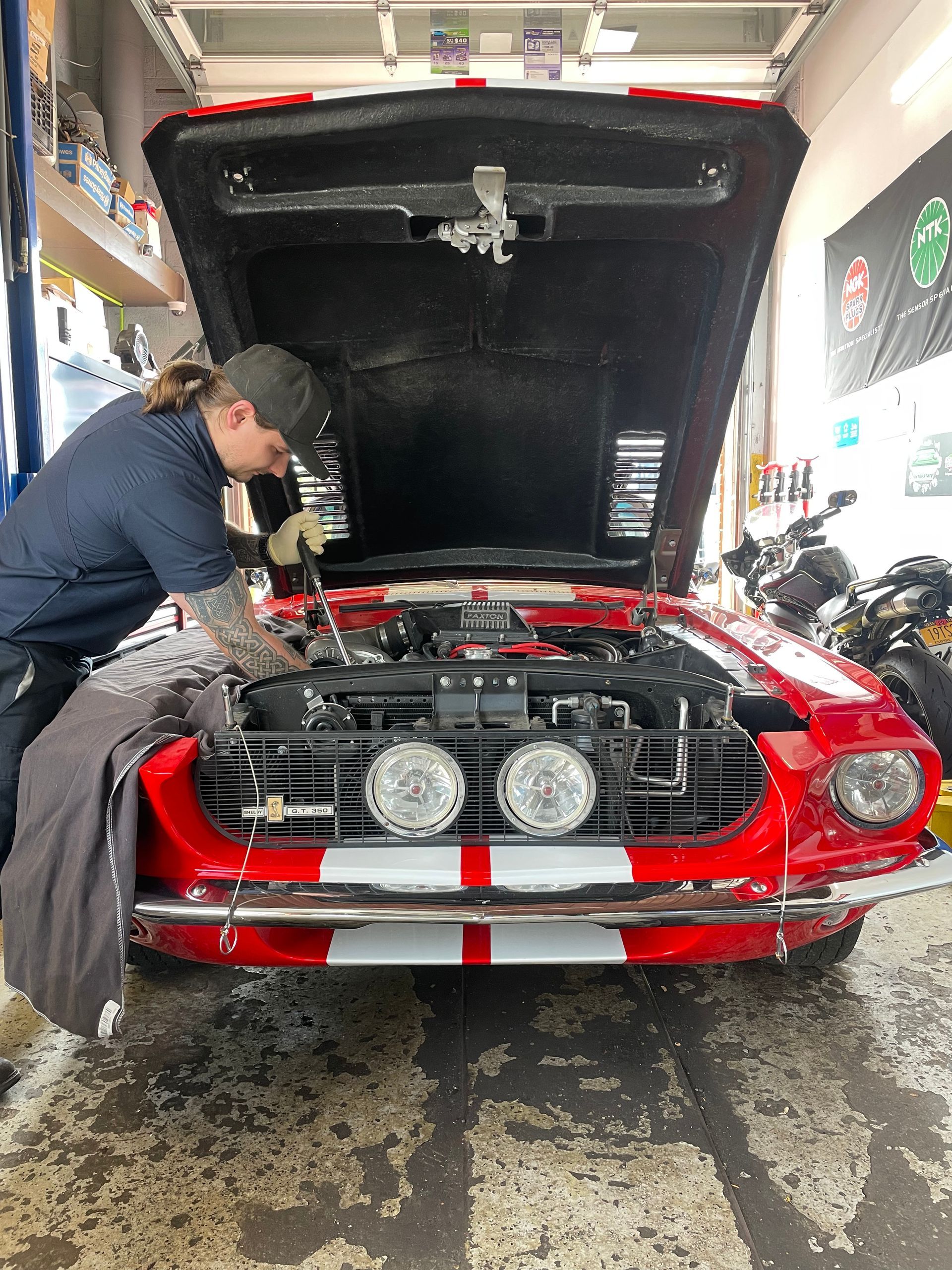 A mechanic works on a red vintage car with a hood open. They wear a cap and are in a garage setting.