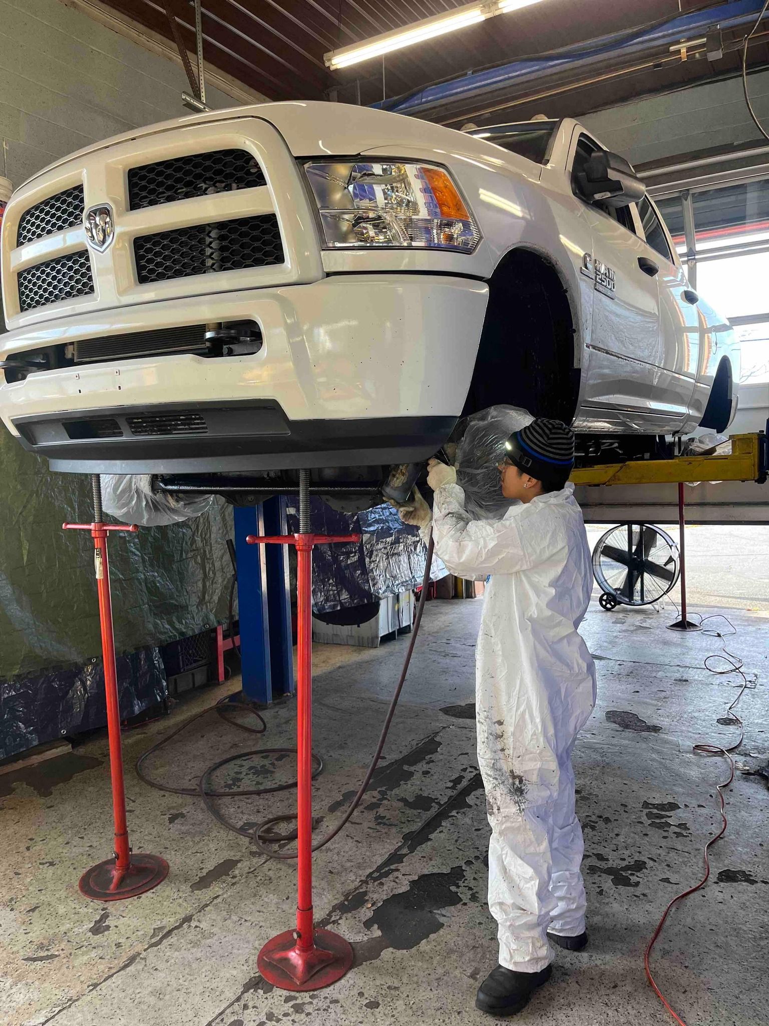 Mechanic in white coveralls working on white truck lifted in a repair shop.