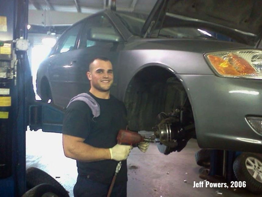 Mechanic smiling at camera while working on a car with the hood open in a garage.