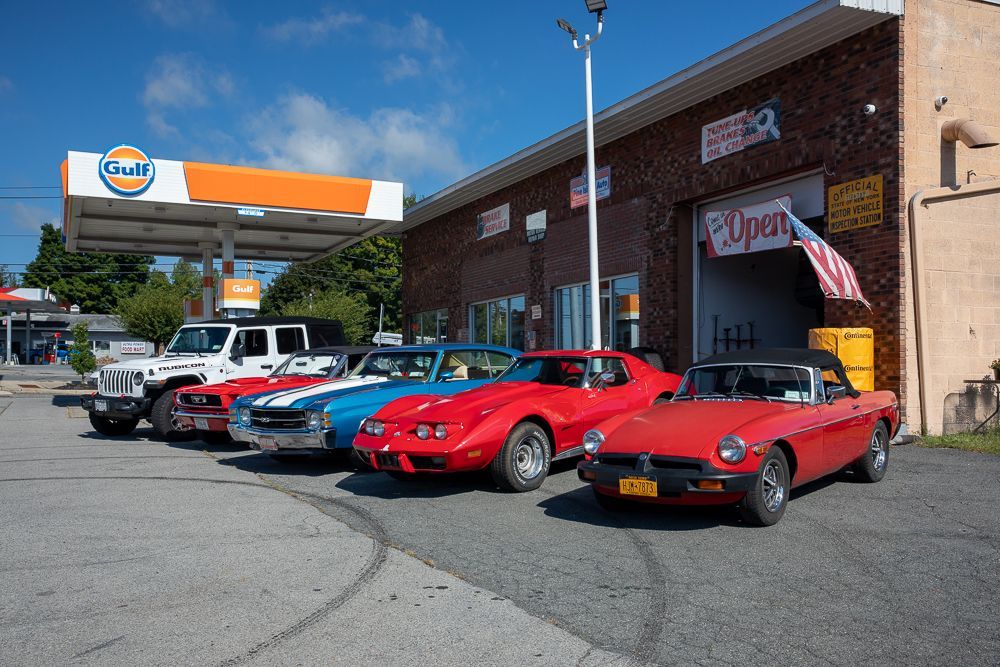 Classic cars parked in front of an auto repair shop with a Gulf gas station in the background on a sunny day.