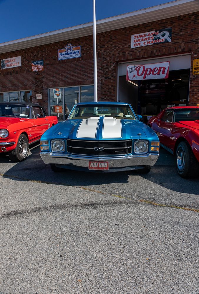 Blue classic car parked outside a brick auto repair shop, flanked by red cars.