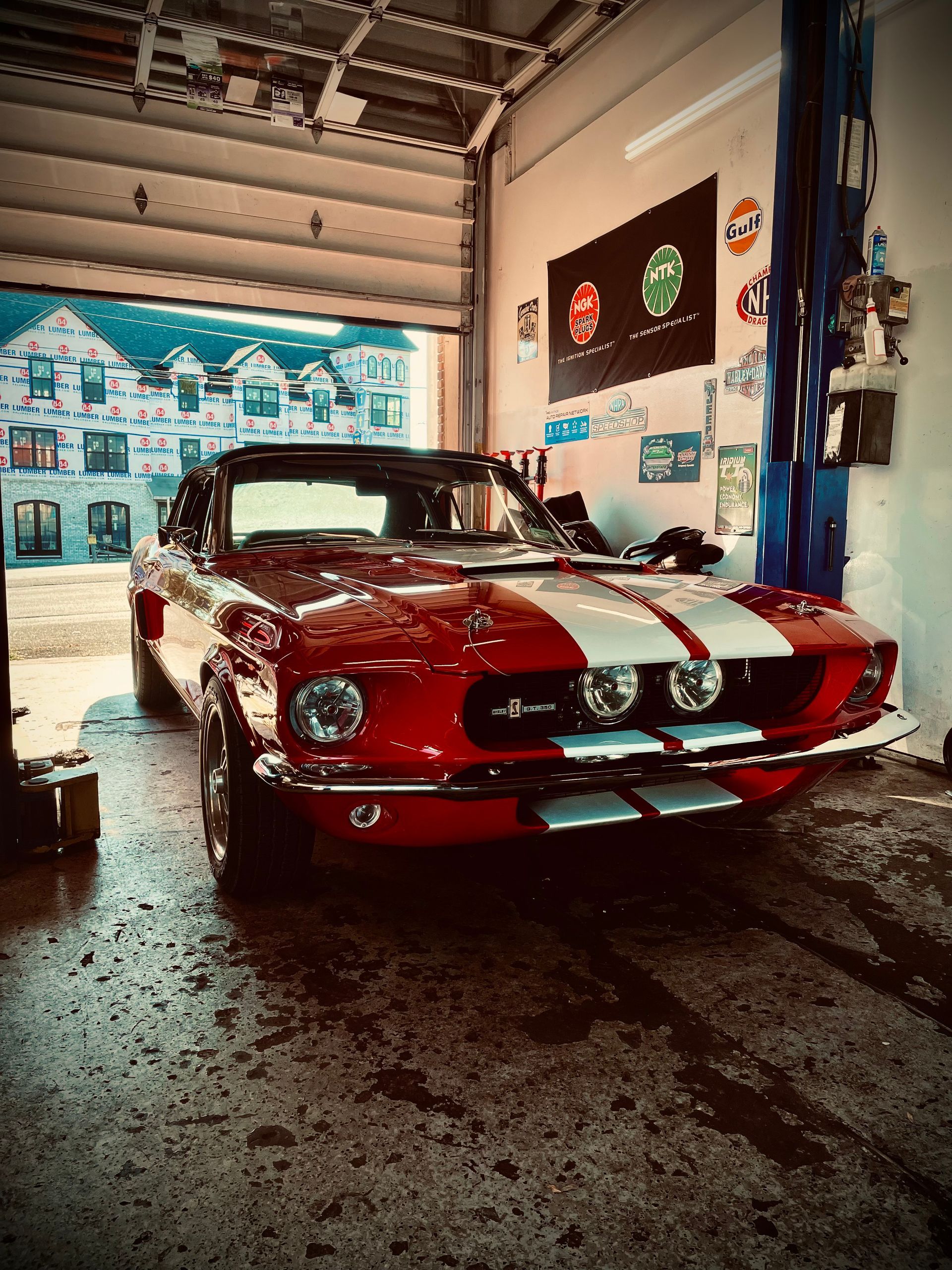 Red Shelby Cobra in a garage, white racing stripes, open garage door with building view.