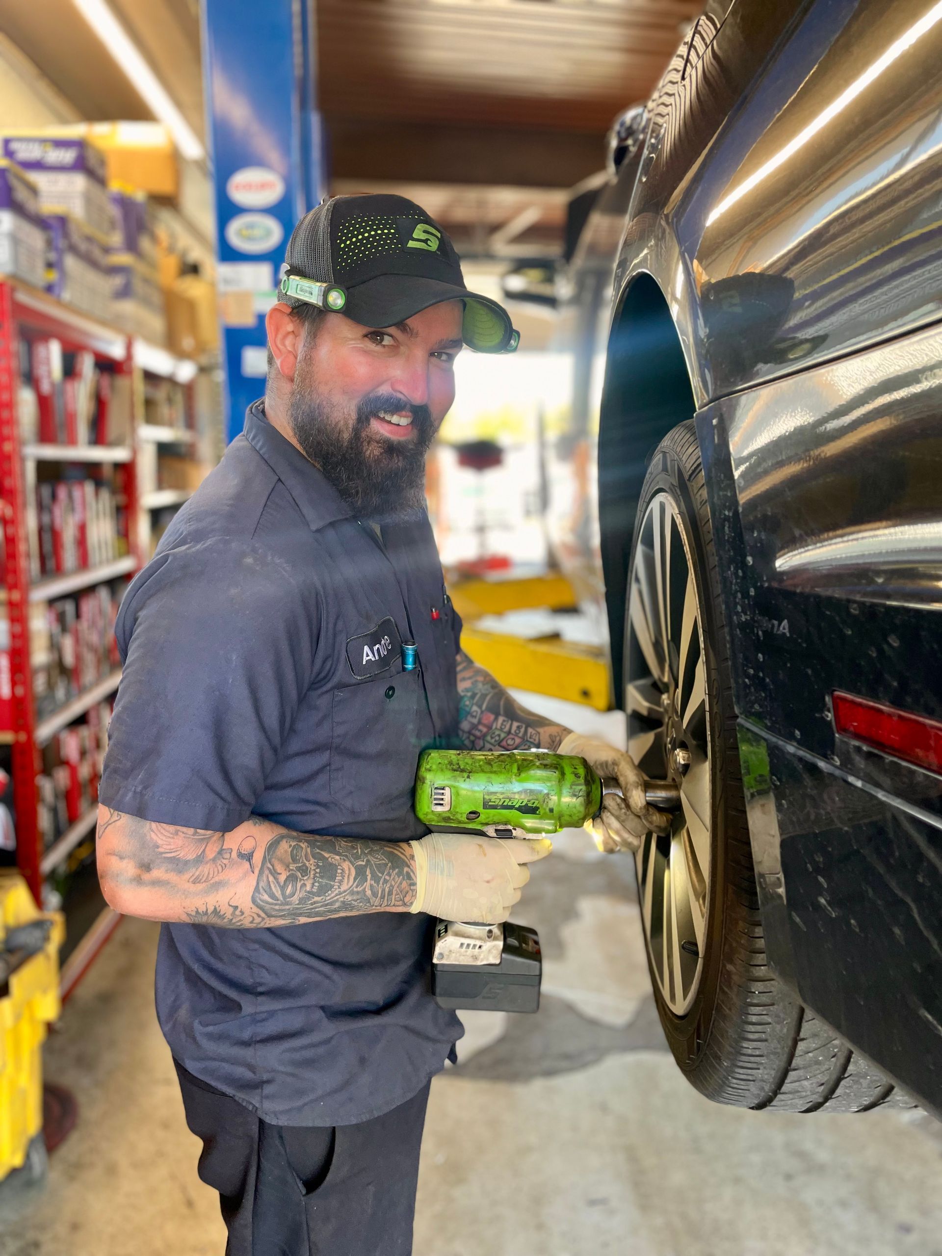Mechanic with tattoos uses power tool to remove wheel from black car in a garage, smiling at the camera.