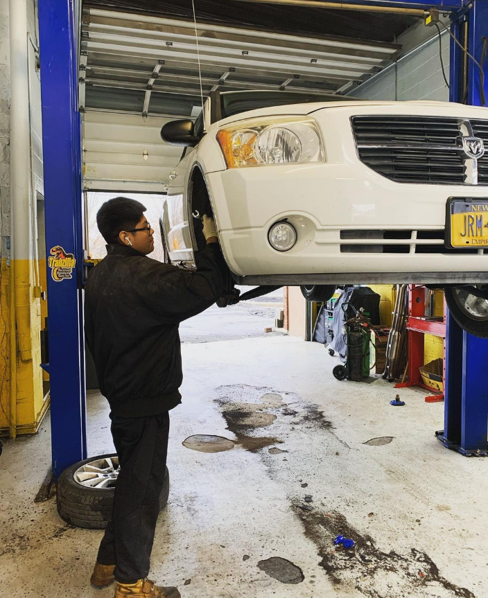 Mechanic working on a white Dodge vehicle elevated on a lift inside a repair shop.
