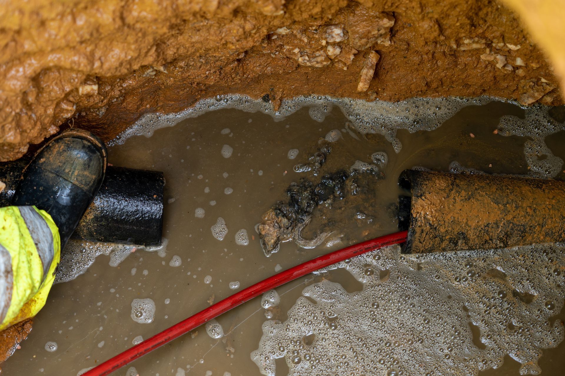 Worker in a trench beside a muddy leaking pipe, with a red hose and standing water.