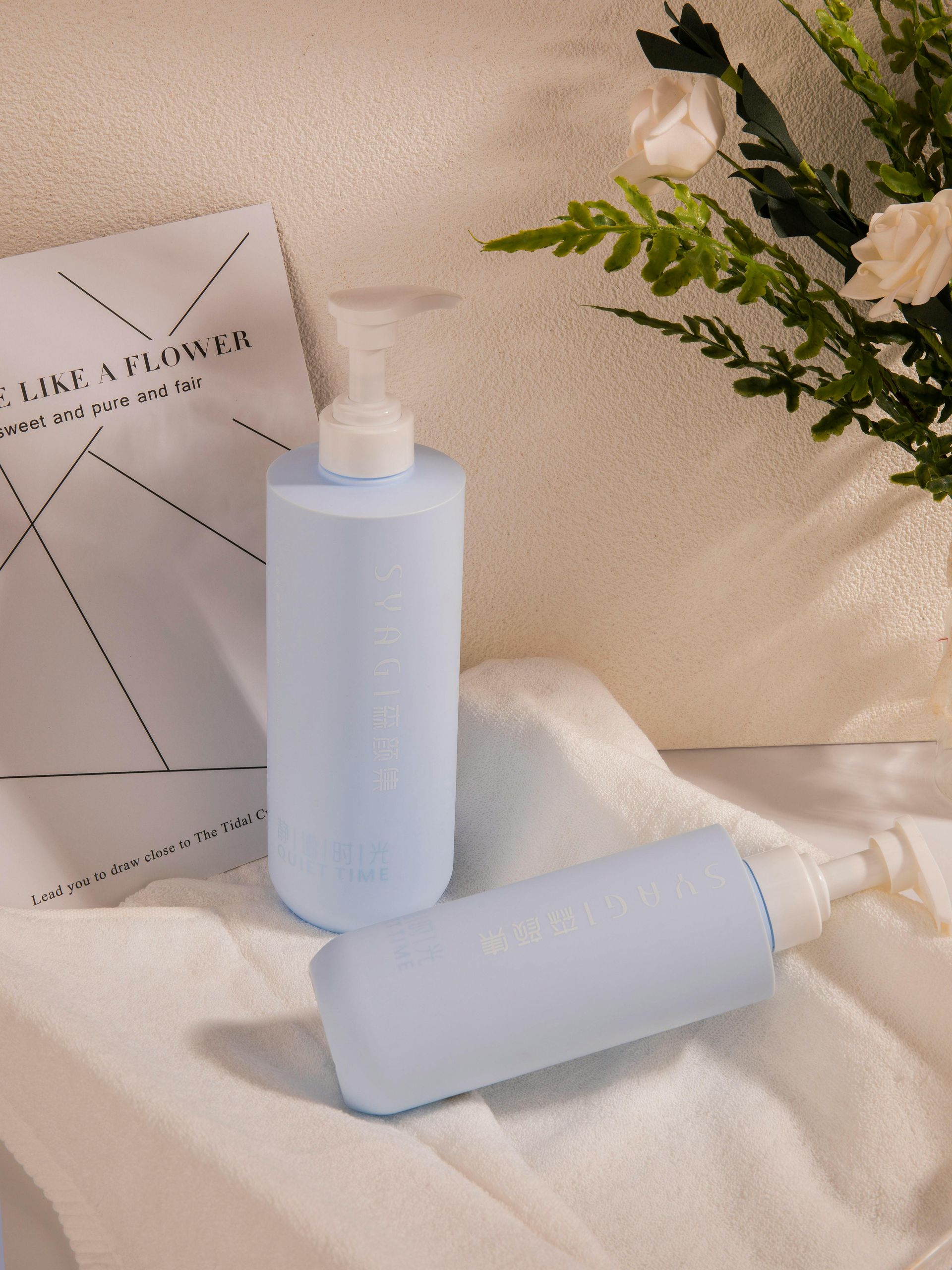 Dos frascos de loción azul claro con dosificadores blancos sobre una toalla blanca. Al fondo, un libro y flores.
