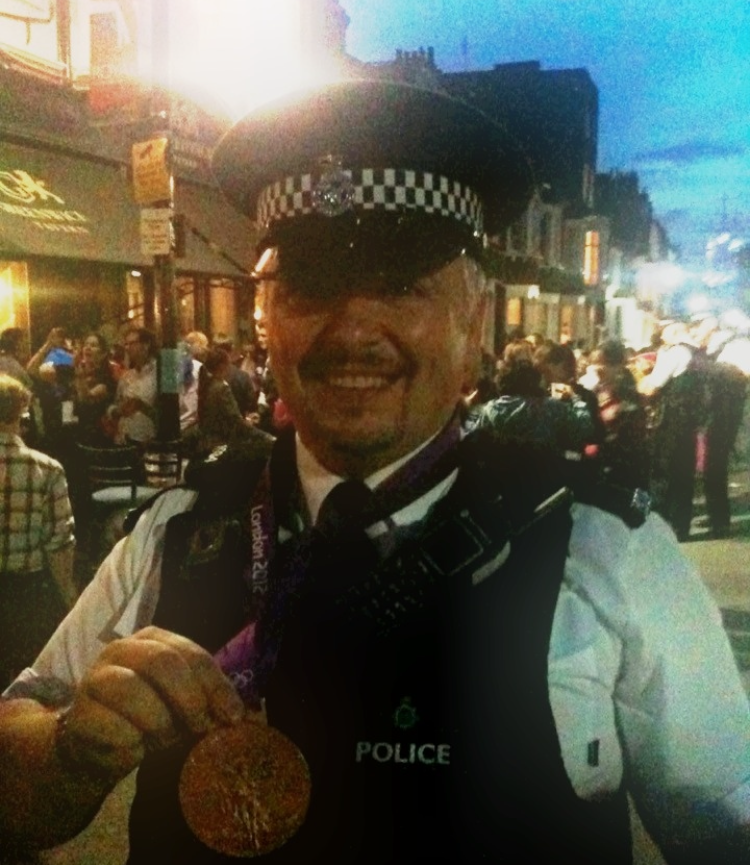 A smiling police officer in uniform holds a small medal, standing on a bustling city street at dusk.
