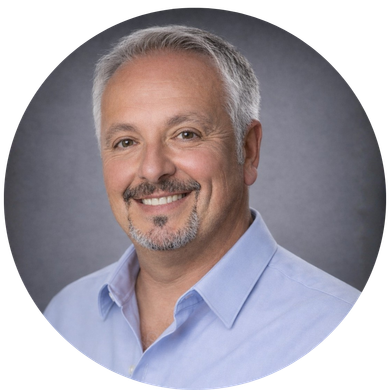 A smiling person with gray hair and a goatee, wearing a light blue button-down shirt against a dark gray background.