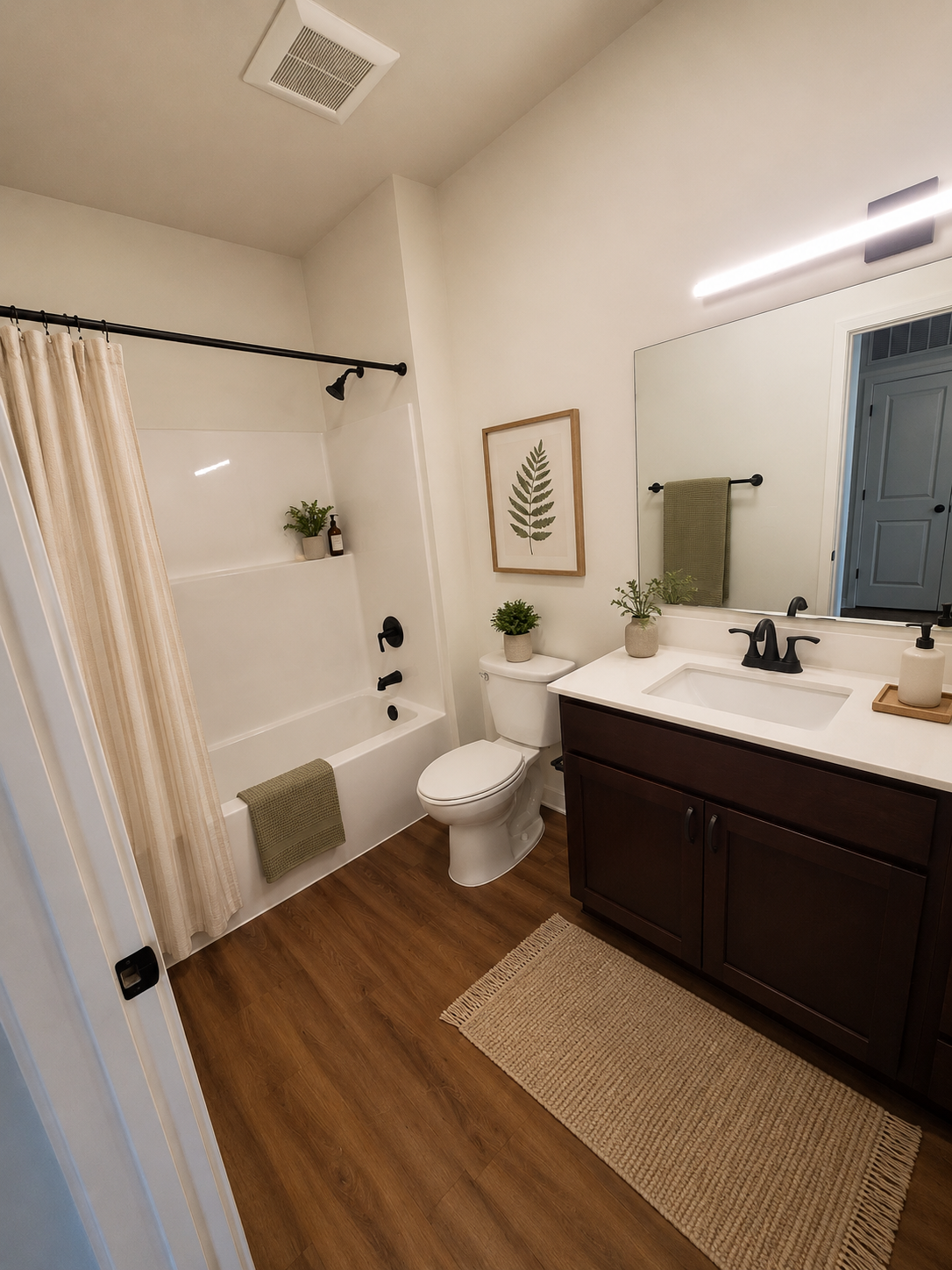Small bathroom with white shower curtain, toilet, dark vanity, sink, mirror, and beige rug on wood-look floor.