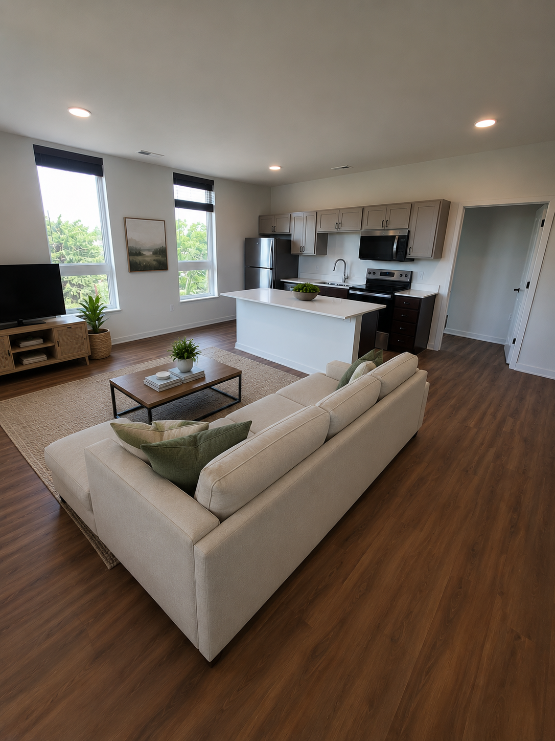 Bright open-plan living room with beige sectional, coffee table, and white kitchen with wood floors.