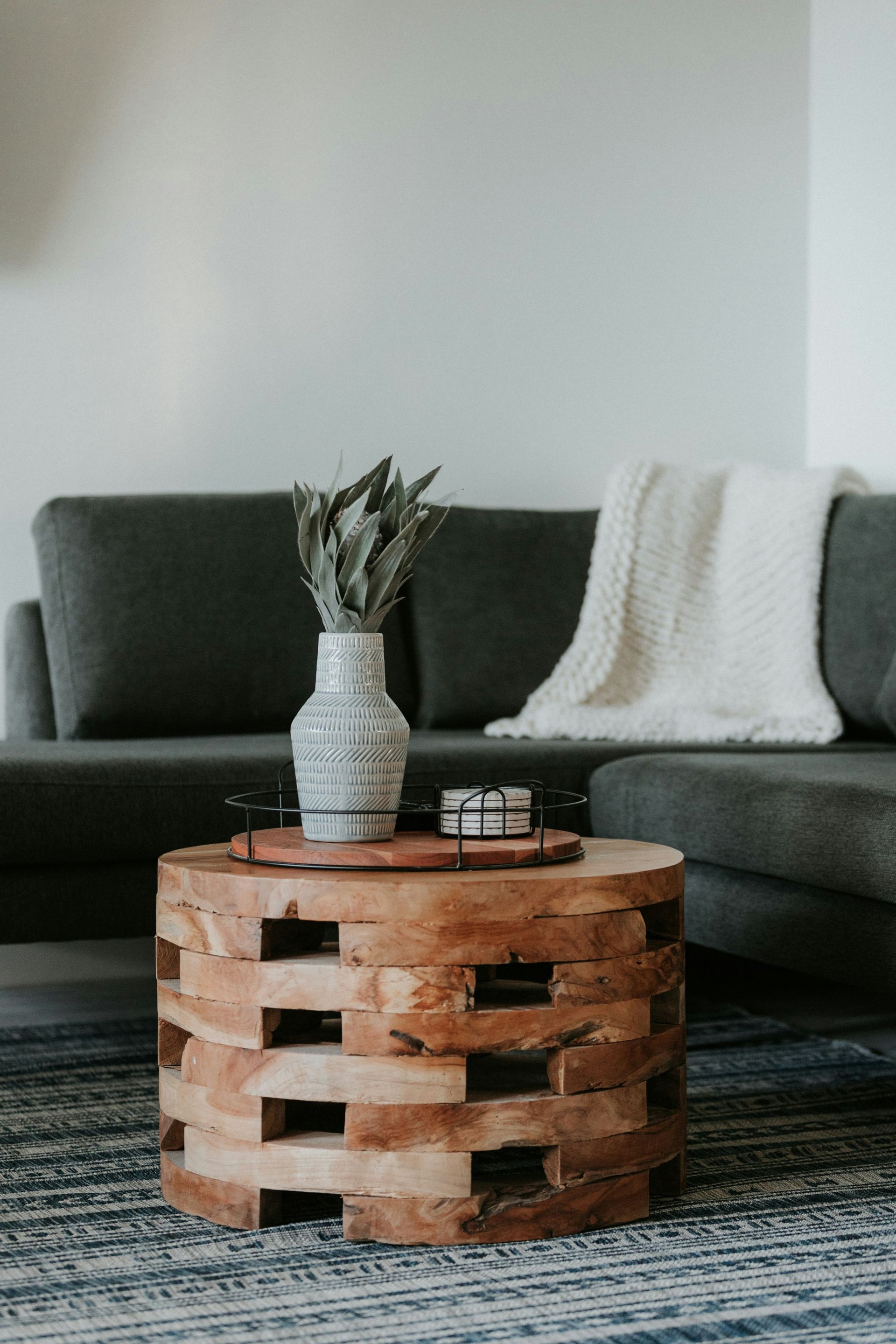Wooden coffee table with vase and sofa. Gray couch with white blanket, blue rug.