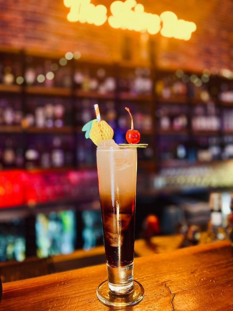 Cocktail in Tall Glass, Layered Dark and Light Liquids, Garnished With Pineapple, Cherry. Bar Background, Brick Wall — The Shire Bar & Grill In Dubbo, NSW