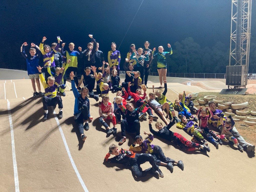 Group of BMX racers on a track at night, raising hands and smiling.