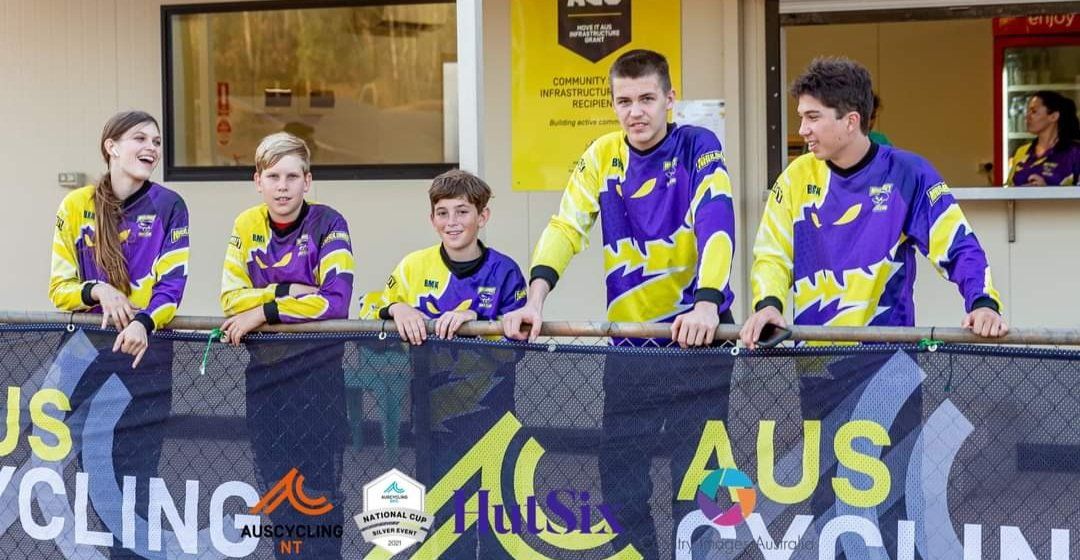 Cyclists in purple/yellow uniforms lean over a fence with