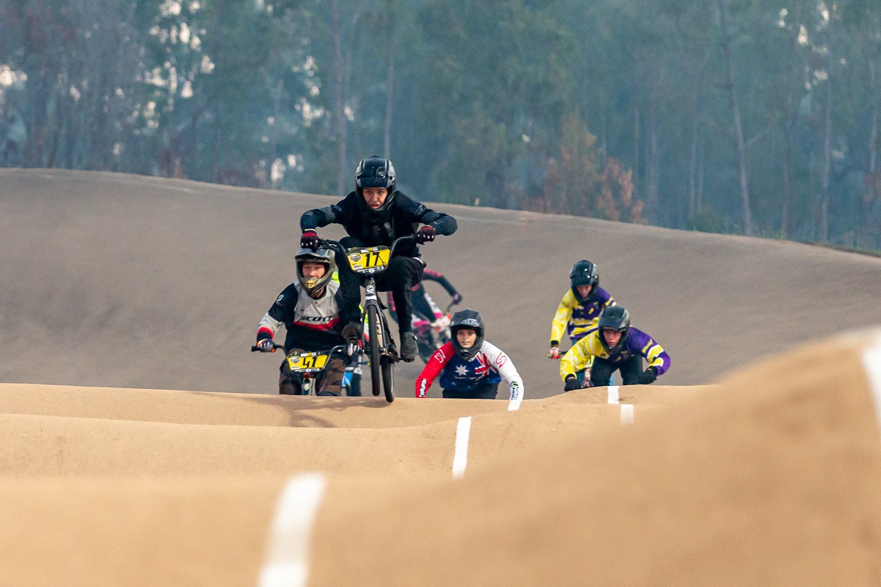 BMX race, riders on dirt track, descending a berm. Leader in black; other riders in various colors.