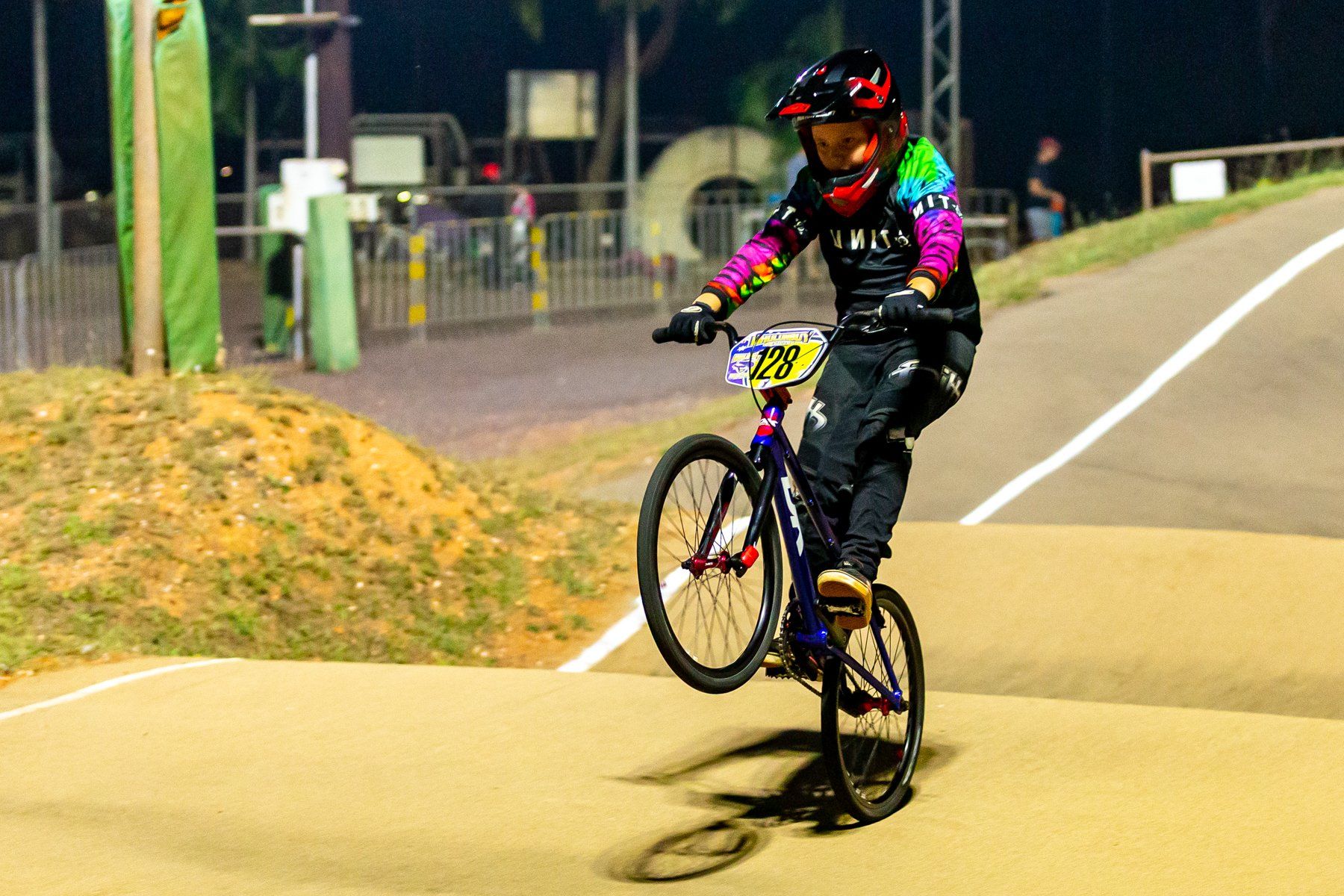 BMX rider on one wheel at a track. They wear a tie-dye shirt, black pants, and a helmet. Nighttime setting.