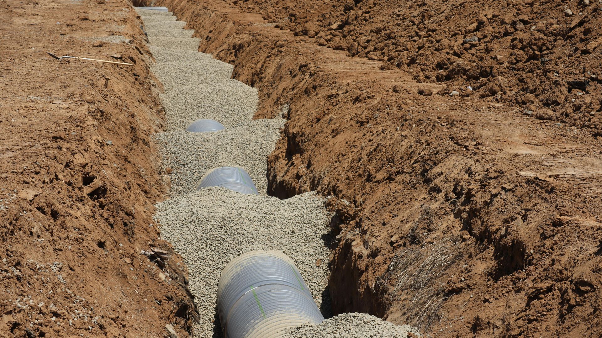 Excavated trench with a large metal pipeline being installed, surrounded by dirt and gravel.