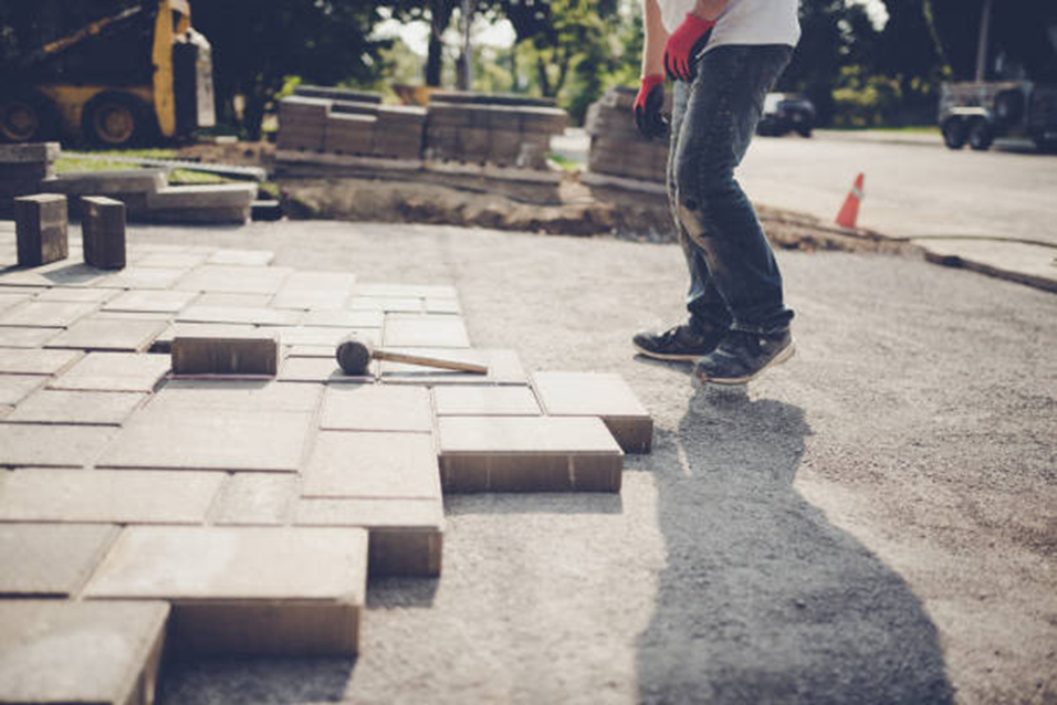 Man laying pavers on a gravel bed; construction site, bricks, hammer, blue jeans, red gloves, sunlight.