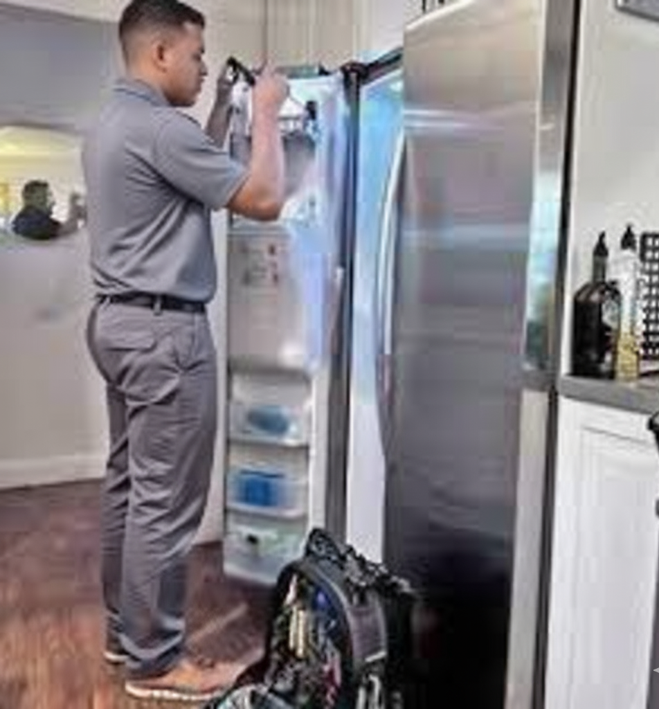 Person in gray uniform repairing a refrigerator, toolbox nearby in a kitchen.