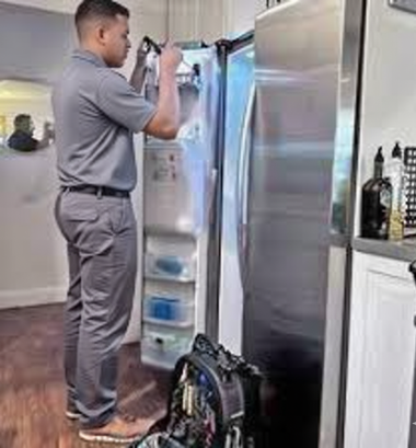Person in gray uniform repairing a refrigerator, toolbox nearby in a kitchen.